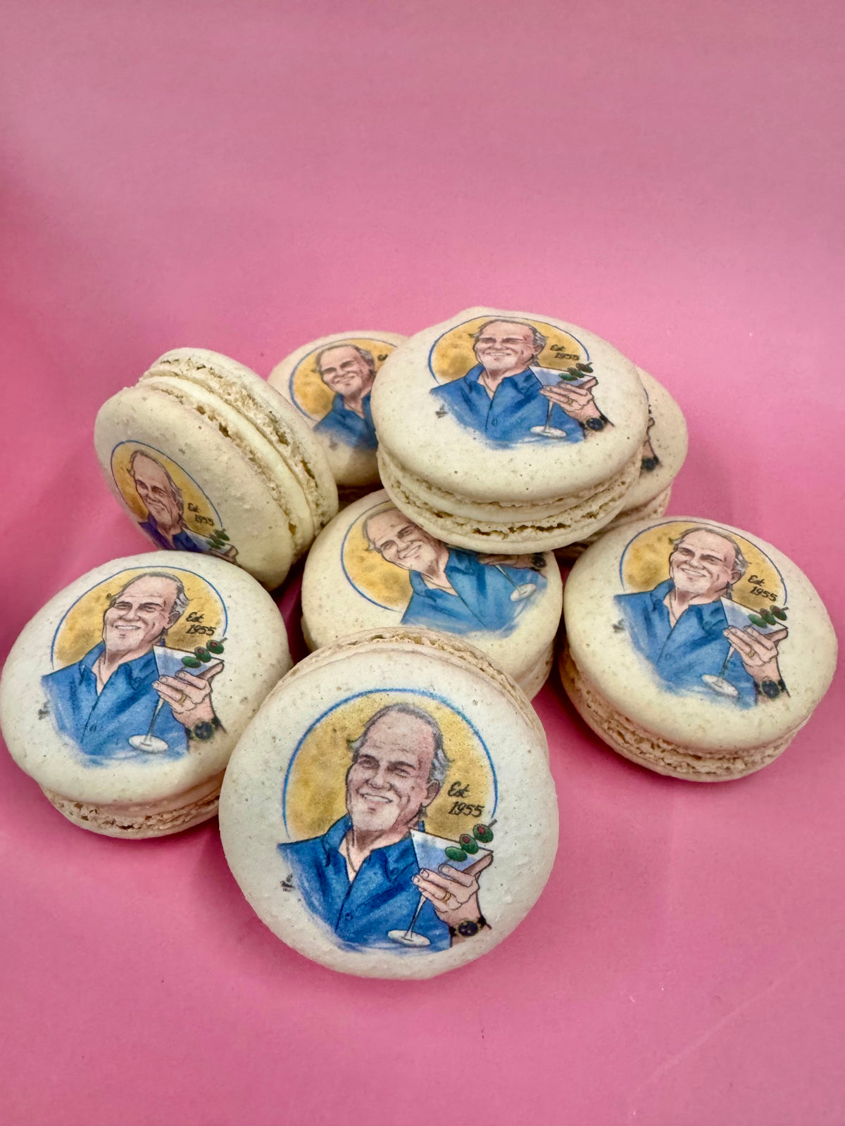 Ivory macarons printed with an illustrated portrait of a smiling man raising a martini, marked Est. 1955, on a pink background