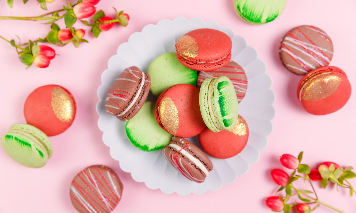 Plate of red, green and red-striped chocolate macarons, some with gold leaf, beside red berry sprigs on pink