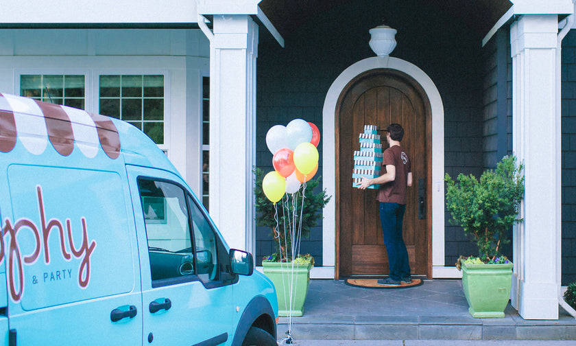 A Trophy driver carries a stack of striped cupcake boxes to a front door, next to a bunch of balloons and the Trophy van.