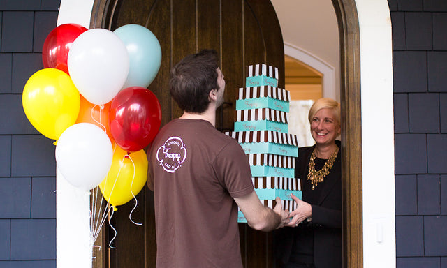 A Trophy driver hands a stack of striped boxes to a smiling woman at her front door, with balloons.