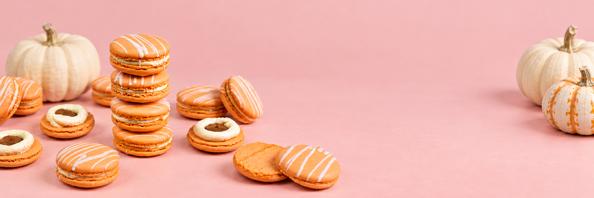 Pumpkin macarons drizzled with white icing, four stacked and the rest scattered among ivory and striped pumpkins on a pink backdrop.