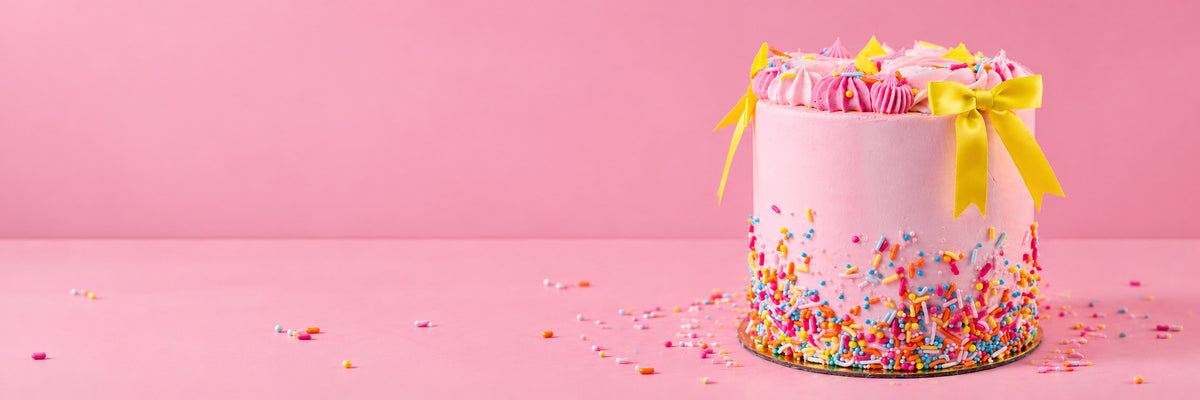 A pink buttercream cake with pink piped swirls, two yellow satin bows and a band of rainbow sprinkles on a gold board, on a pink table scattered with sprinkles.