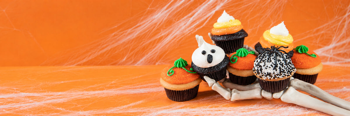 Halloween mini cupcakes with ghost, candy corn, pumpkin and spider decorations arranged on a skeleton hand, against an orange backdrop with cobwebs.