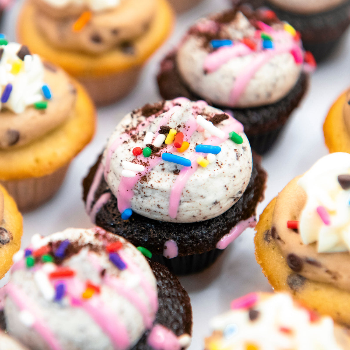 Close-up of mini cupcakes with cookies-and-cream frosting, pink drizzle and rainbow sprinkles beside chocolate chip cookie minis