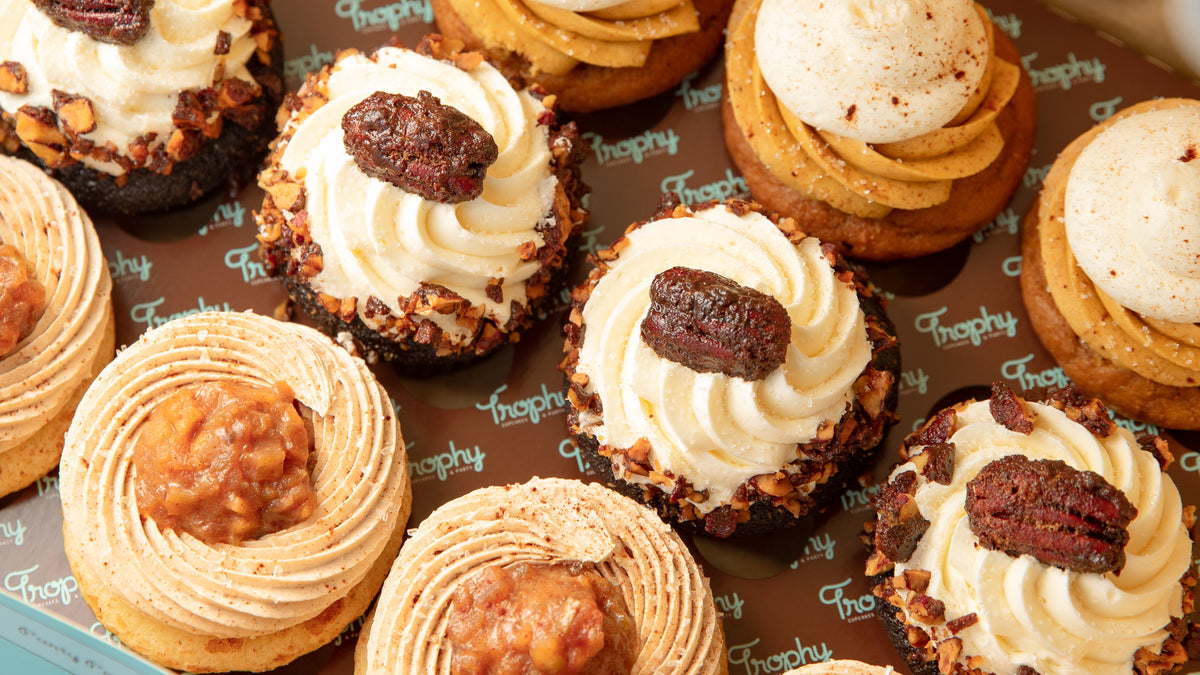Box of fall cupcakes: white frosting with candied pecans, pumpkin-colored frosting, and spiced frosting topped with fruit filling