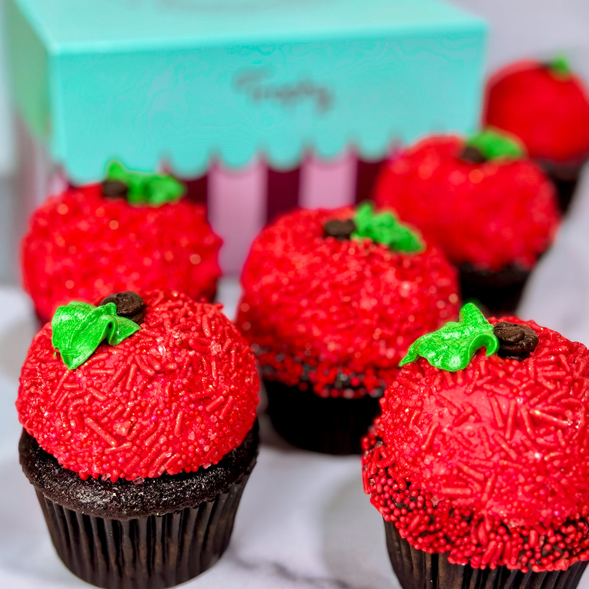 Red sprinkle-covered cupcakes shaped like apples with green leaf toppers in front of a teal box