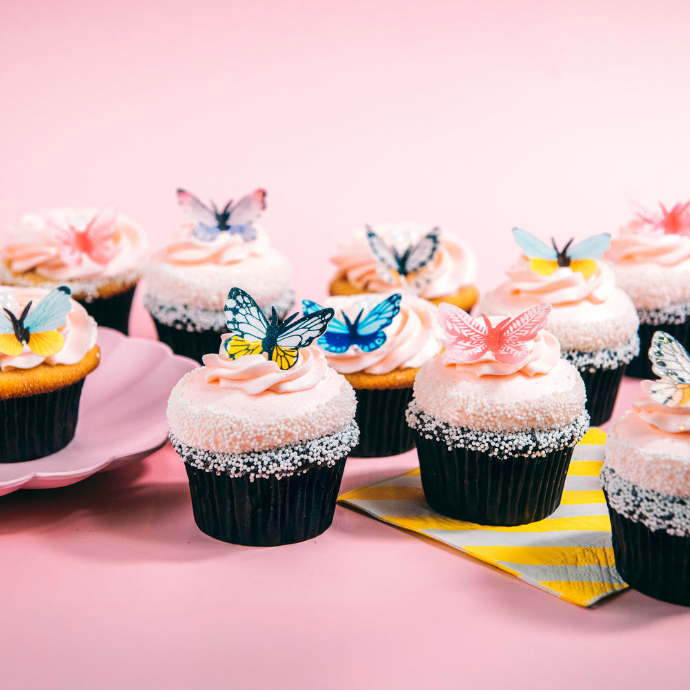 Pink-frosted cupcakes topped with edible butterflies on a pink background beside a yellow striped napkin