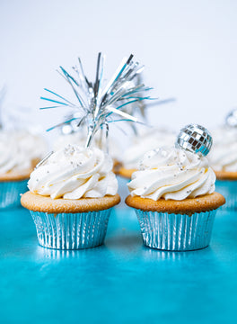 Vanilla cupcakes with white buttercream, a silver tinsel pick and mirror ball toppers on a teal surface