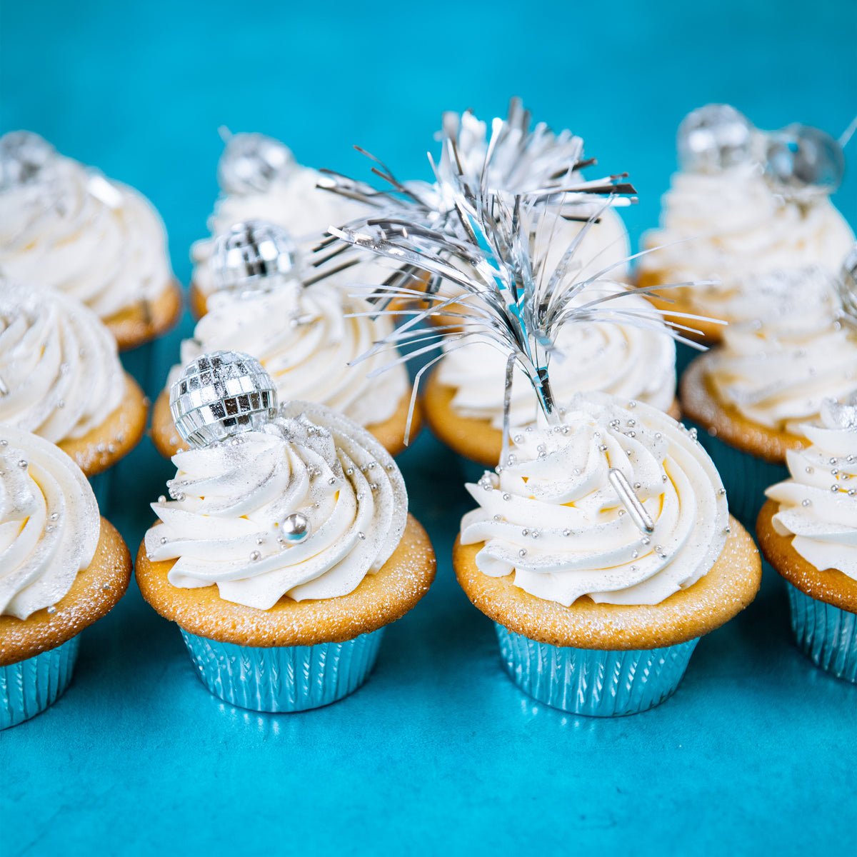 Rows of vanilla cupcakes with white buttercream, silver tinsel picks and mirror ball toppers on teal