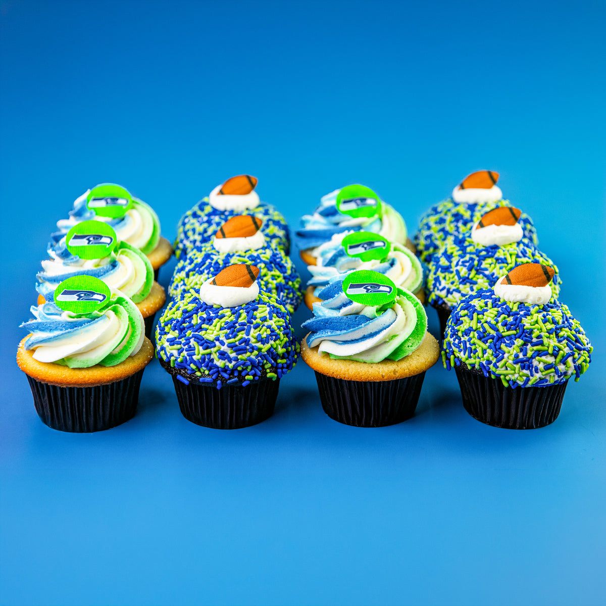 Dozen cupcakes in rows on blue: blue and green sprinkle cupcakes with football toppers and blue, green and white swirled cupcakes with green team logo toppers
