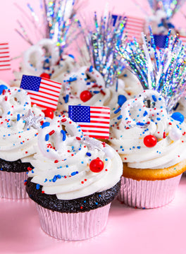 Cupcakes with white frosting, red, white and blue sprinkles, small American flags and silver tinsel toppers on pink