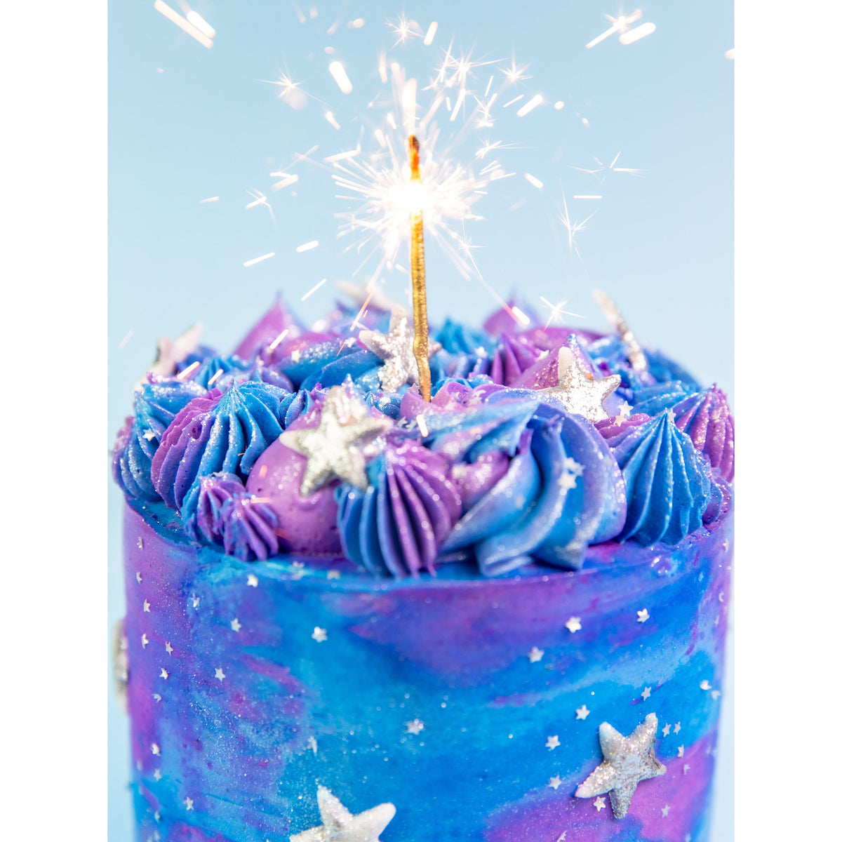 Galaxy Cake with a lit sparkler candle, blue and purple buttercream swirls and silver stars